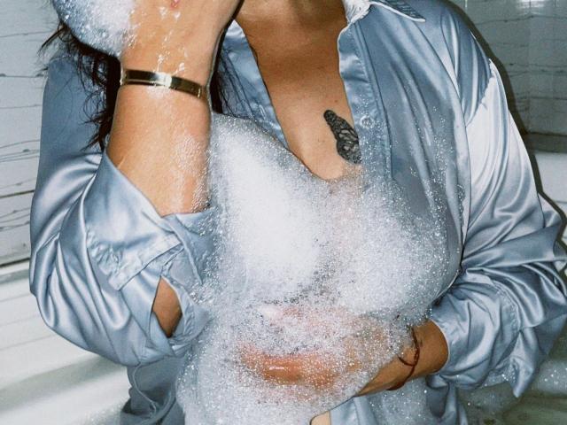 LorenaLoveS in a bubble-filled bath, wearing a shiny, blue satin shirt, hands covering her tits, a small tattoo on her chest, gold bracelet, soap suds on her face.