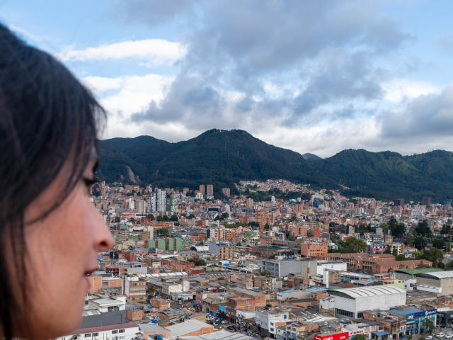 JainaRogers' face in the foreground, with dark hair and brown skin, looking at a city with mountains and cloudy sky in the background. City has red and white buildings.