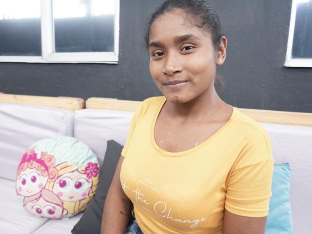 MakenaAnderson with dark skin, curly hair, yellow shirt, small tits, smiling, sitting on a white couch with colorful pillow, blue pillow, and black wall behind.