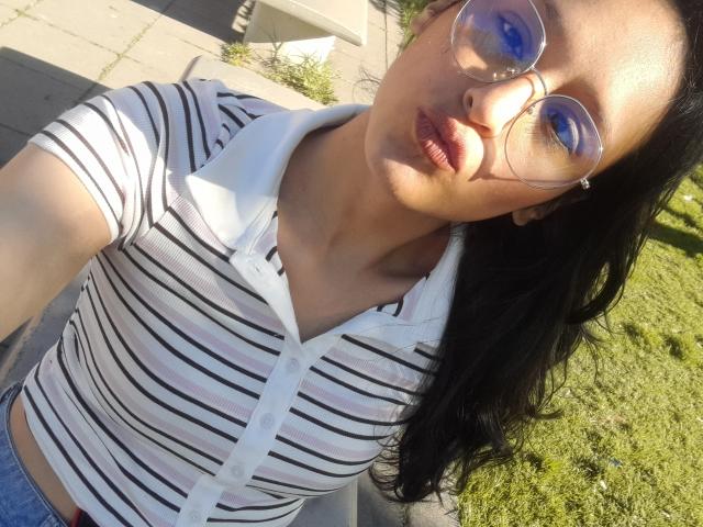 WorthyOfBdsm has dark hair, wears striped shirt, white collar, blue eyes reflected in glasses, pouty lips, and is outside in sunlight. Boobs visible under shirt. Grass in background.