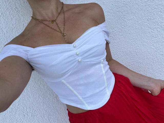 DarkKrokus-hot wearing a white off-shoulder top with buttons, red skirt, and gold necklace, showing small tits and a slim waist. White textured wall in background.