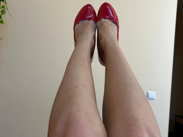 MoonLiza's legs are crossed at the ankles, wearing shiny red high heels. Her pale skin is visible, and she's sitting against a beige wall.