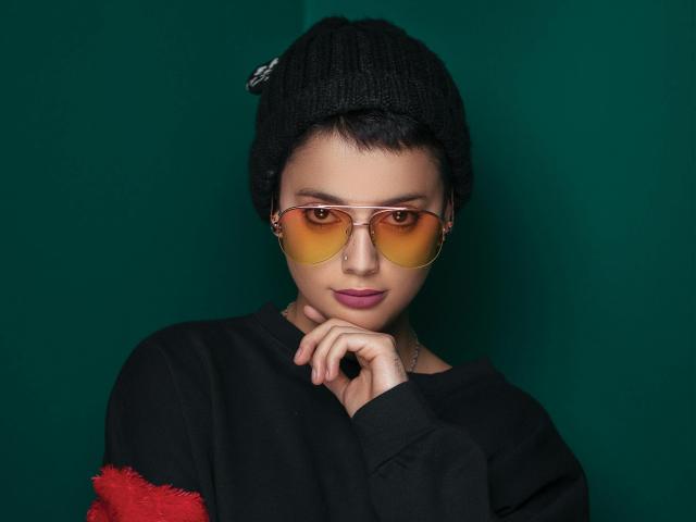 CamilaRosx with short black hair, wearing a black beanie, orange-tinted aviator sunglasses, and a black sweater with a red fluffy patch, touching her chin.