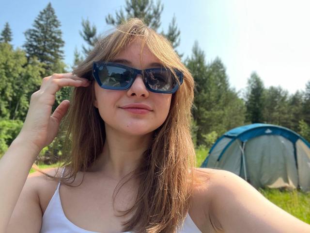 MayxFlower, con i capelli castani, gli occhiali da sole blu e la canotta bianca, che si tocca i capelli, in piedi di fronte a una tenda blu, sorridendo, in una foresta soleggiata..