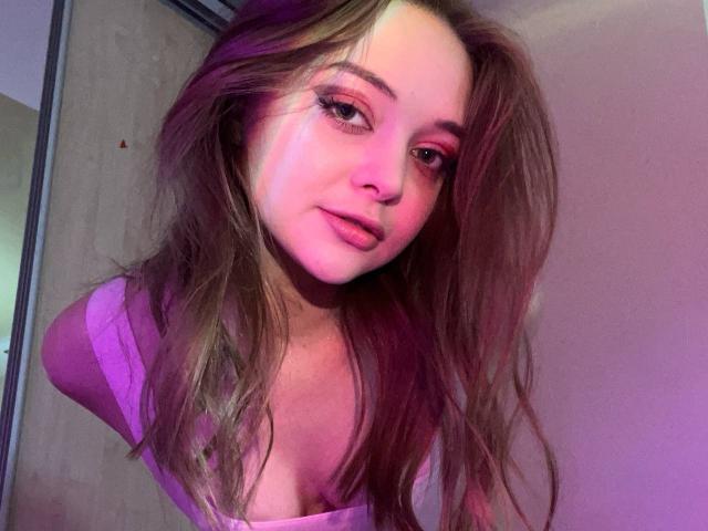MayxFlower hat langes, braunes Haar, pinken Lidschatten und ein pinkes Oberteil, mit einem leichten Lächeln und einem süßen Gesicht, auf einem Selfie, das in violettem Licht aufgenommen wurde. Ihre Brüste sind dezent..
