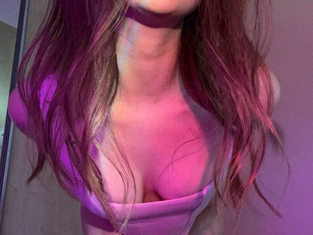Les seins roses de MayxFlower sont visibles sous un haut violet à décolleté bas, avec de longs cheveux ondulés brun-rosés et un collier violet, sur une photo prise légèrement en biais, montrant le haut du corps..
