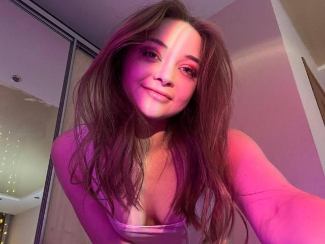 MayxFlower con una luce rosa e viola sul viso, sorridente, con lunghi capelli castani, indossa una maglietta rosa che mette in mostra il décolleté, mentre si fa un selfie leggermente inclinato..