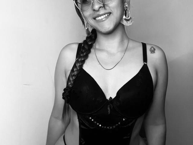 Black and white photo of AlannaN with glasses, braided hair, smiling, wearing a black lace bra, necklace, and earrings, showing small tattoo on right shoulder, and a cute face.