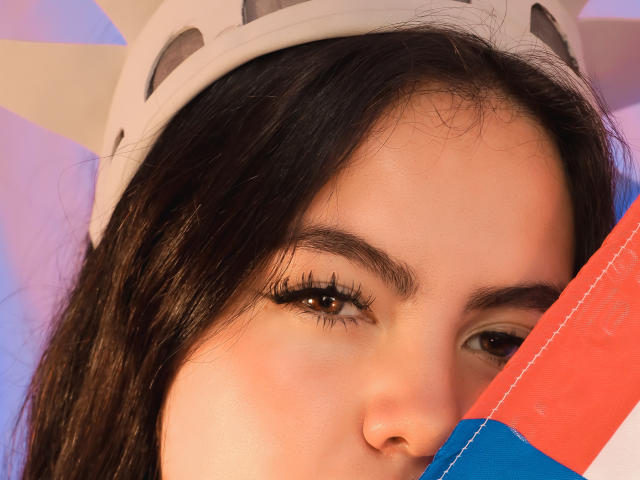 ZoeQuinn's face with long dark hair, wearing a white helmet, brown eyes with thick black eyelashes, partial red and blue striped object in front. Soft, natural skin.