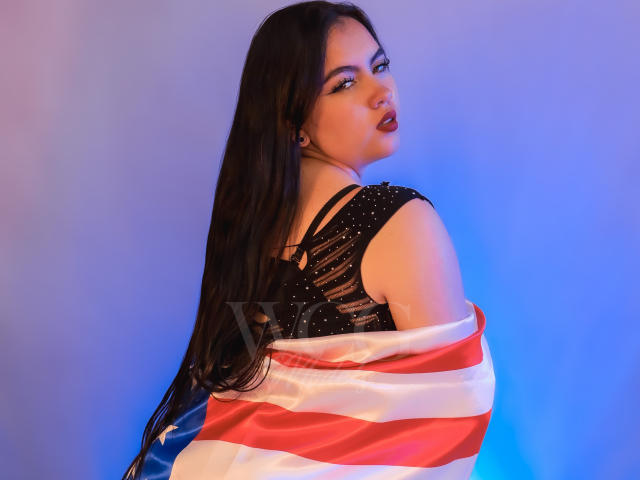 ZoeQuinn with long black hair, wearing a black sparkly top, and an American flag draped over her shoulders, looking back with red lips, blue eye makeup, and a sexy smile.