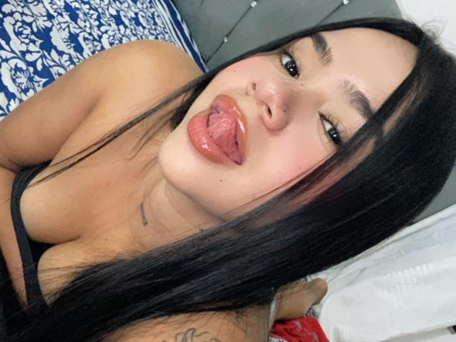 DaphnePeython has long black hair, dark eyes, and is sticking her shiny pink tongue out. She's wearing a black top, showing a tattoo on her right shoulder.