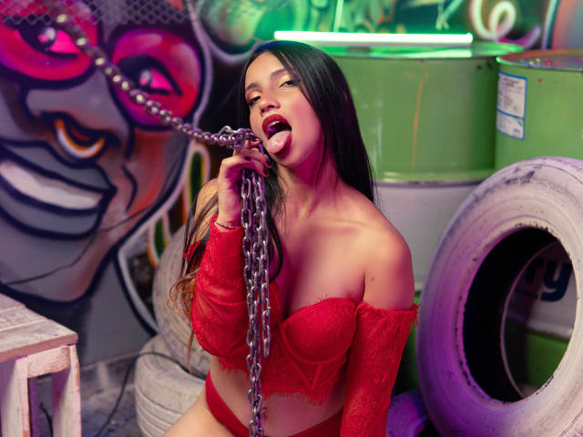 MiluMilan with long black hair, red lace off-shoulder bra, and matching panties, licking a metal chain, showing her tongue and teeth. She's in a gritty, colorful urban setting.