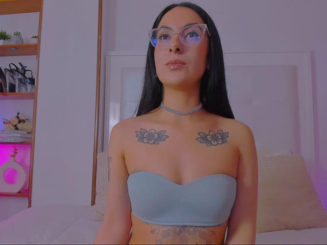 SarahLuna with long black hair, glasses, and a strapless blue top showing her tits and tattoos of flowers on her chest. She's wearing a choker and has a tattoo below.