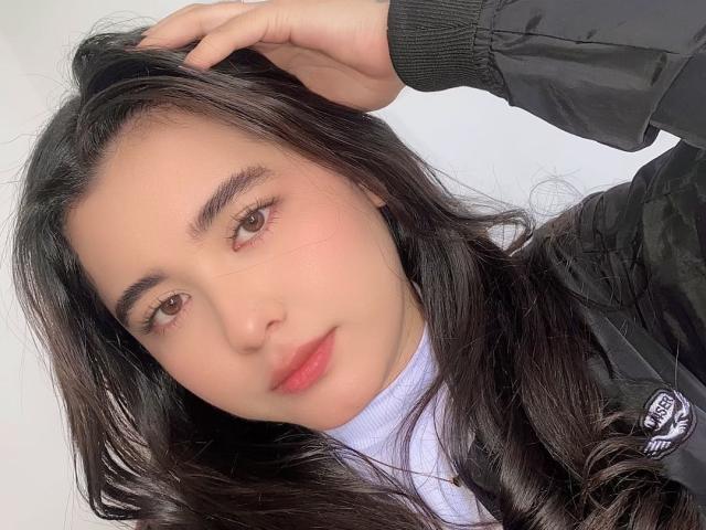 LianaBernocchi with long black hair, fair skin, brown eyes, red lips, wearing a black jacket, hand in hair, slight smile, white turtleneck, soft light, casual selfie.