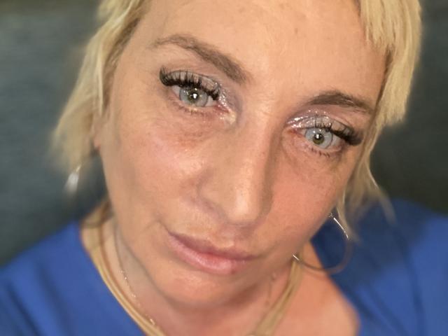 LunaSensualX has blonde hair, green eyes, and wears blue. She's looking at the camera with a slight smile, wearing hoop earrings and a gold necklace. Her face is slightly wrinkled.
