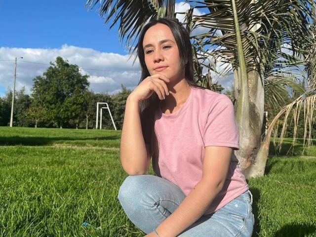 Lila69Hot, agachada, camiseta rosa, jeans azuis, mão no queixo, palmeira, grama verde, céu azul claro, nuvens brancas, sorriso suave, cabelo castanho claro, fofa, relaxada..