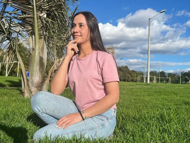 Lila69Hot, com cabelo escuro, camisa rosa e jeans azuis, sentada na grama, com a mão perto da boca, com uma expressão pensativa, em um dia ensolarado, com uma palmeira ao fundo e um céu azul claro..