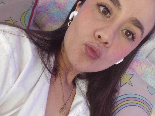 Lila69Hot tiene ojos marrones, labios rosados y gruesos, y una camisa blanca, lleva auriculares blancos, tiene el pelo largo y oscuro, y un collar dorado, y está haciendo una mueca..