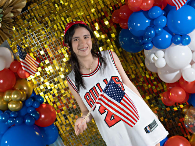 ValerieBanks with dark hair, red headband, white "Bulls" jersey, holding American flag, smiling, surrounded by red, blue, and white balloons, gold sequin background, in festive setting.