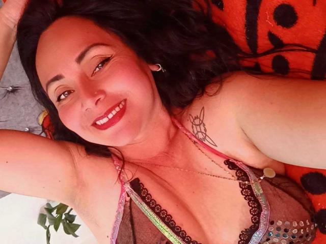 Zaria69 with dark hair, smiling, wearing a sheer, lace-trimmed, pink and black bra, showing her tits and a tattoo on her right shoulder, laying on an orange blanket.