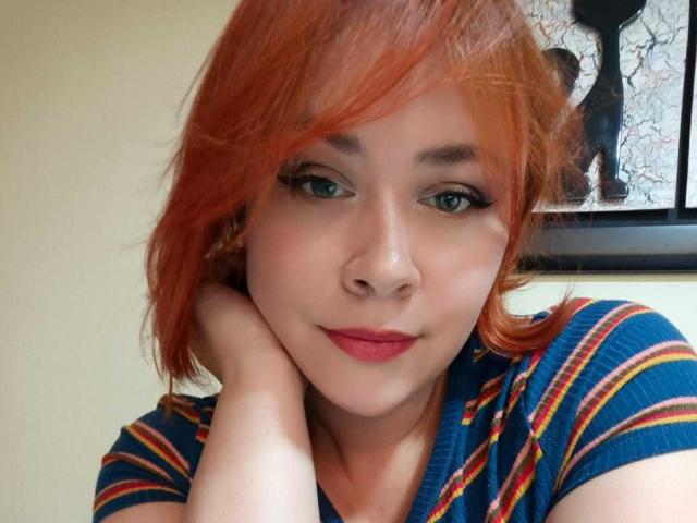 MaddyBlooms with red hair, green eyes, and red lipstick, wearing a blue, striped shirt, resting her chin on her hand, smiling slightly. Her tits are small, visible under shirt.