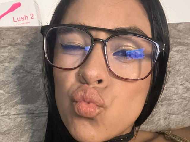 StellaBeckker with puffy pink lips, black-framed glasses, and a nose ring, making a pouty face, wearing a black choker, and reflecting blue lights in her eyes.