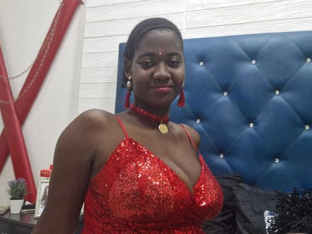 VioletaWal with dark skin, wearing a red sequined dress, choker, and earrings, sitting in front of a blue tufted headboard, smiling at the camera, showing her tits.