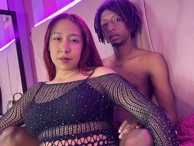 LaurenMike with red hair, black mesh top, and small tits, sits in front of a shirtless black dude with dreads and a tattoo on his chest. Both look serious.