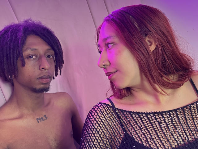 LaurenMike with brown dreads, nose ring, and chest tattoo, stares at LaurenMike with red hair, nose ring, and fishnet top, purple light on right. LaurenMike's tits visible.