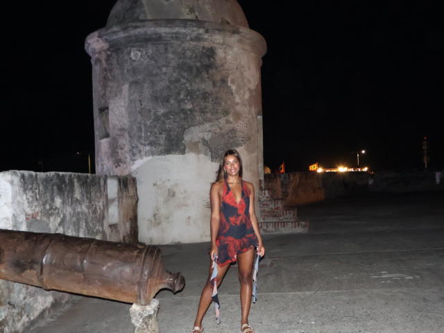 EmmaLatin with dark skin and long brown hair, wearing a red and black dress, stands in front of a large, ancient stone structure at night, with a cannon on the left.