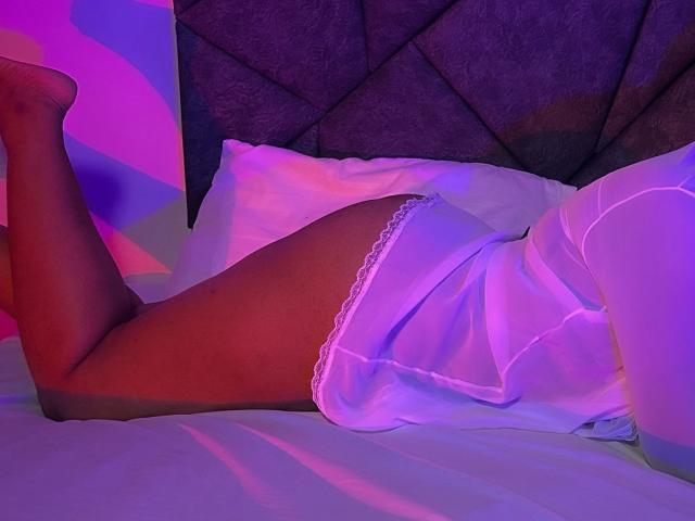 Kaassandraa lying on a bed, legs bent, wearing white lace-trimmed panties and a white dress, illuminated by purple and pink lights, with her ass and thighs visible.