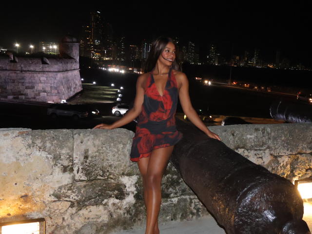 EmmaLatin with dark skin, long brown hair, and red floral dress, leaning on old cannon, city lights in background, night, sexy, slim, small tits, bare legs, stone wall.