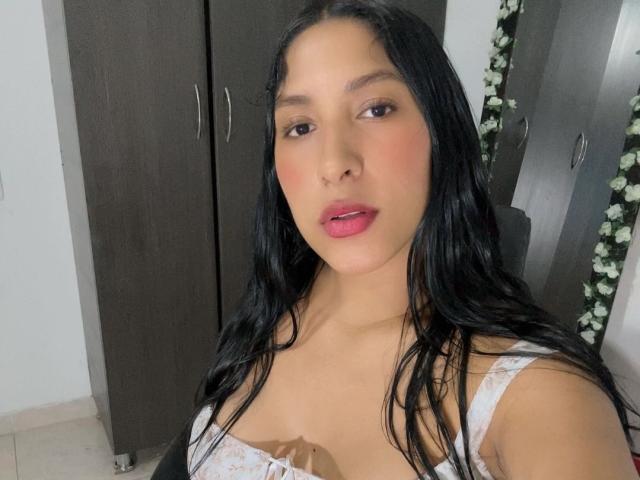 MadisonReyes has long black hair, pink lips, and a light tan skin tone. She's wearing a white, floral-patterned top, showing her small, perky tits. She's holding a brown object.