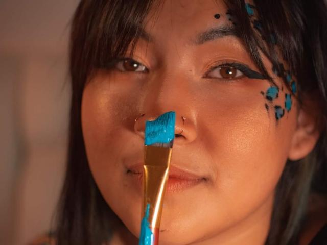 KionI's face with blue paint on nose, cheek, and eyebrow, holding a gold paintbrush with blue paint, brown eyes, black hair, nose ring, slight smile.