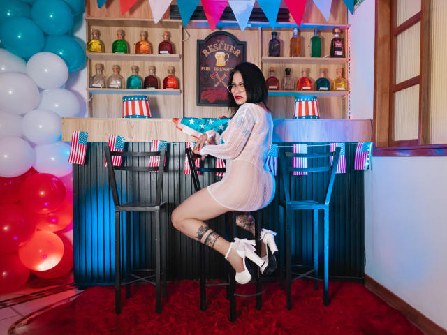 Donna Jones, avec ses cheveux noirs, portant une robe blanche transparente, est assise sur un tabouret de bar, montrant ses fesses et ses talons hauts blancs, tenant un objet bleu avec un motif d'étoiles, fond en bois d'un bar..