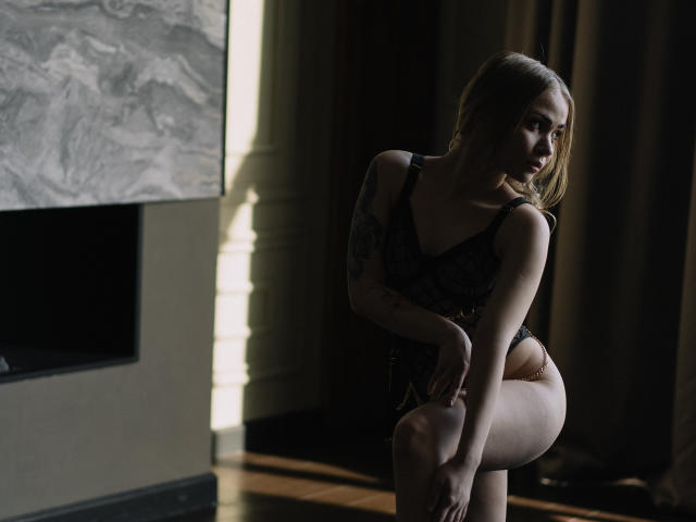 AmelyCollie with small tits, tattoo on right arm, wearing black lace lingerie, sits in shadow, light on left, brown hair, pale skin, hands on knee, dark brown curtain behind.