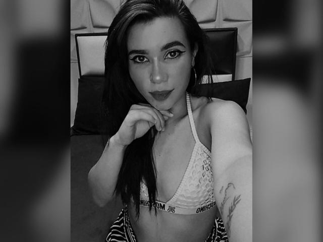 Charlott Jones tiene el cabello oscuro, lleva un top de encaje, mira hacia arriba con un delineador de ojos llamativo, la mano derecha cerca de la barbilla, tiene un tatuaje pequeño en el brazo izquierdo, es un selfie en blanco y negro..
