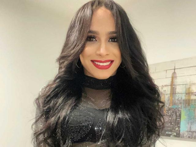 SophiaJimenezX with long, wavy black hair, red lipstick, and a black, sparkly, sheer top, smiling at the camera, showing her shiny, shiny tits.