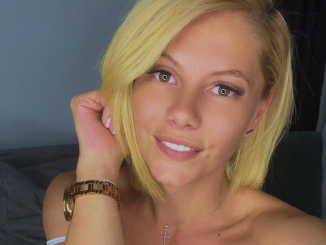TanyaGrey with blonde hair, smiling, blue eyes, and a gold bracelet, showing a cute face and small cross necklace, has a small beauty mark on her right cheek.