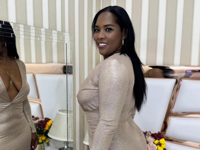 MeridaRoa with dark skin, black hair, and large tits in a shimmering beige dress, smiling at the camera. Reflection in mirror shows her cleavage. Bedroom setting.