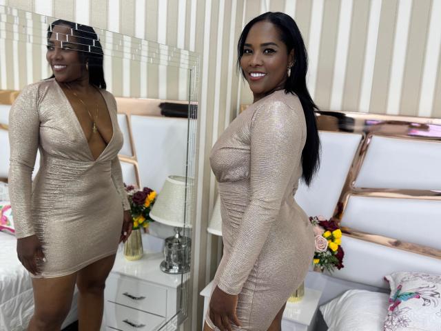 MeridaRoa with dark skin, long black hair, and big ass, wearing a shiny, tight, beige dress with deep cleavage, smiling at her reflection in a mirror.