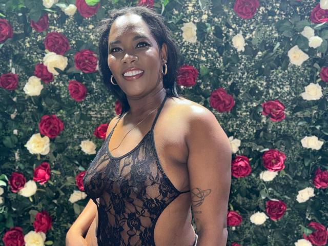 MeridaRoa with dark skin, wearing a sheer black lace bodysuit, showing small tits, smiling, black hair, hoop earrings, tattoo on right arm, standing in front of red roses.