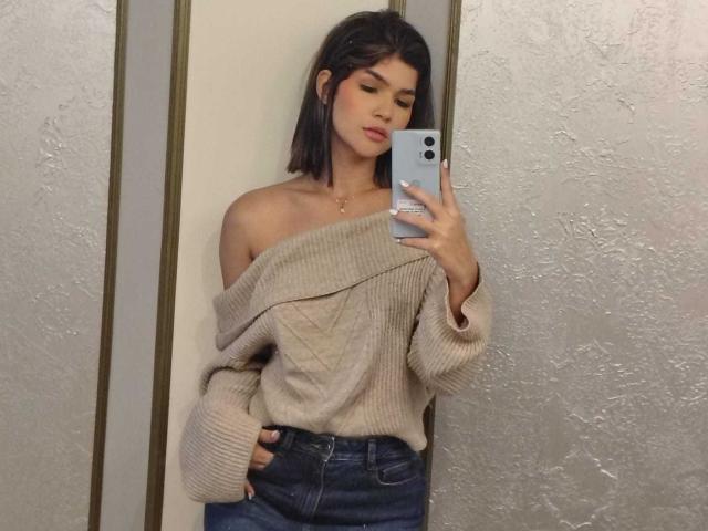 ScarlletZhen with a cute face, dark bob, off-shoulder beige knit, and blue jeans, taking a mirror selfie with her phone, hand in pocket, looking down.