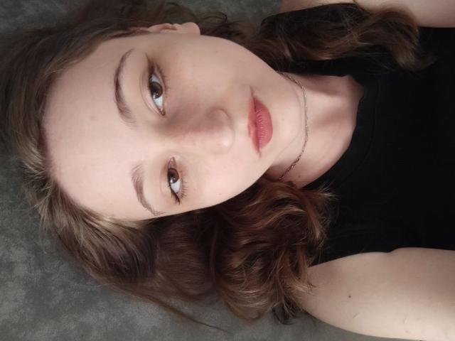 TwinkyKiss has wavy brown hair, light skin, and is wearing a black top with a thin necklace, lying down with a subtle smile, showing her brown eyes and pink lips.