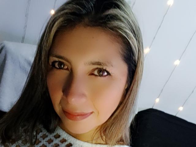 MichelleDumont with light brown eyes, a subtle smile, and blonde-streaked brown hair, wearing a white, leopard-print sweater, in a well-lit room with string lights.