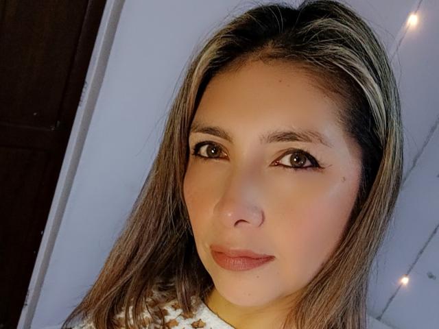 MichelleDumont has brown eyes, light brown skin, and straight brown hair with blonde highlights. She's wearing a white top with a pattern, and has a small mole on her right cheek.
