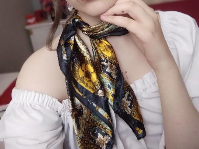 TwinkyKiss with a colorful snake-pattern scarf, white off-shoulder top, and pale skin, touching her chin, showing her bare shoulders and upper chest. Her hand is near her face.