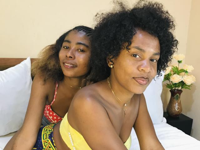 Duas mulheres negras com cabelo cacheado, JulianaSilvie na frente com uma blusa amarela e seios pequenos, JulianaSilvie atrás com uma blusa vermelha com bolinhas e seios médios, sorrindo na cama..