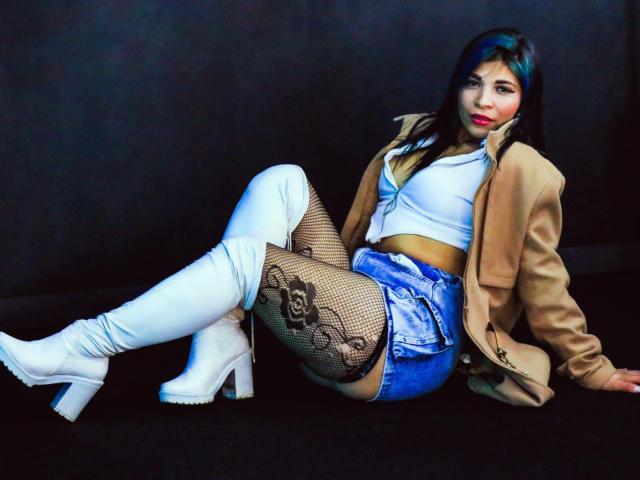 KatalinaMachado with blue denim shorts, white crop top, brown jacket, white knee boots, fishnet stockings, and dark hair, sitting on black floor, legs crossed, looking at camera.