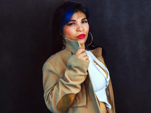 KatalinaMachado with blue highlights, wearing a tan jacket, white top, and red lipstick, touching her chin with a ring on her finger, large hoop earrings, dark background.